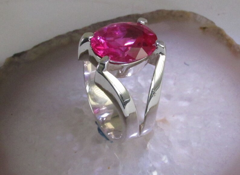 May include: A silver ring featuring a large, faceted, pear-shaped, hot pink gemstone. The ring's band has a unique, open design with curved, flowing lines that cradle the stone. The ring is set against a light background.