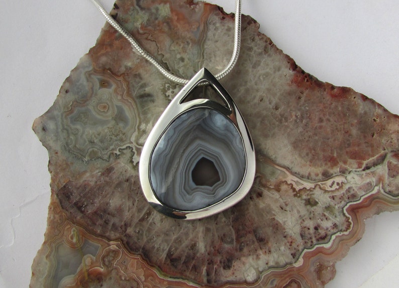 May include: A teardrop-shaped pendant with a polished silver frame and a gray and white agate stone center. The pendant hangs from a silver chain. The stone has concentric bands of gray and white. The pendant is displayed on a stone surface.