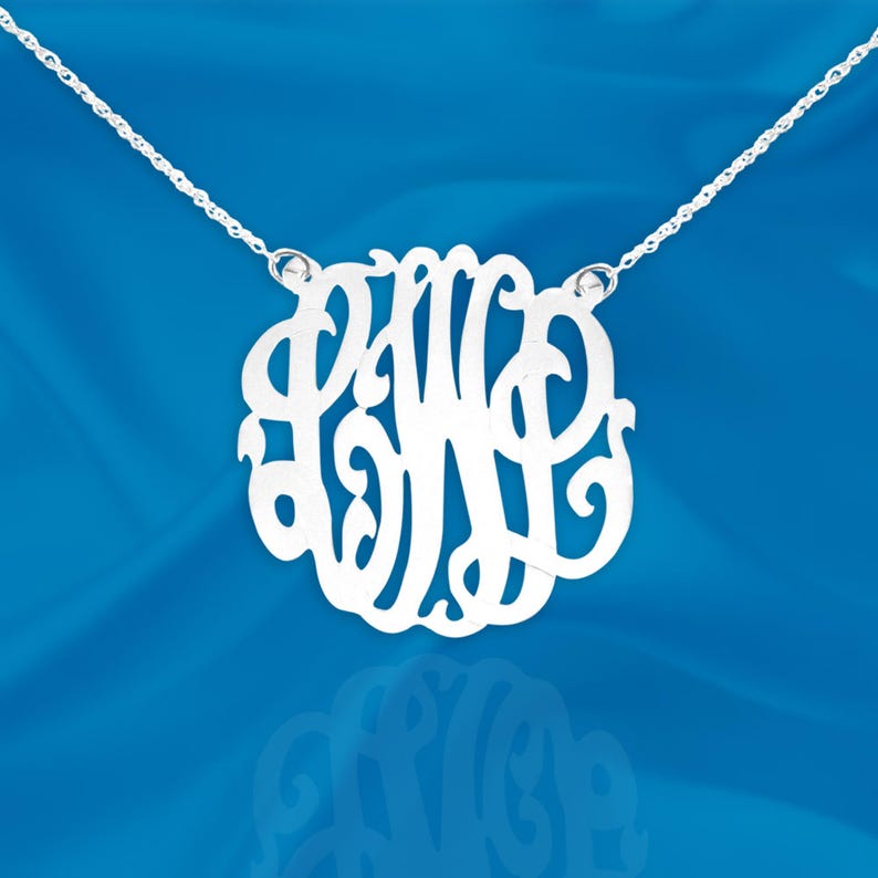 May include: A silver necklace with a delicate chain and a large, openwork monogram pendant. The pendant features the letters "PWB" in a stylized script font.