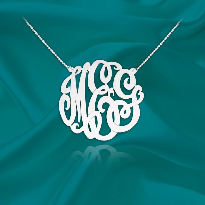May include: A silver necklace with a large, circular monogram pendant featuring the letters "MGC" in a cursive script.
