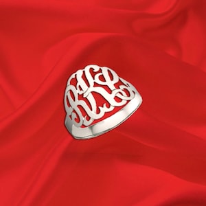 May include: A silver ring with an ornate, openwork design featuring three intertwined initials. The ring is set against a vibrant red background, creating a striking contrast. The ring's polished surface reflects light, highlighting its intricate details.