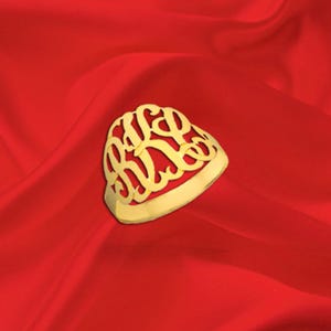 May include: A gold-colored ring with an ornate monogram design, set against a vibrant red background. The ring's monogram features intertwined letters, creating a personalized and elegant accessory. The ring is likely made of gold or a gold-toned metal.