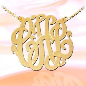 May include: Gold necklace with a large, ornate monogram pendant. The pendant features three letters intertwined in a decorative script.