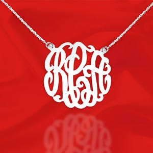 May include: A silver necklace with a large, round pendant featuring a monogram design. The pendant is made of a single piece of metal and has a delicate, intricate design.