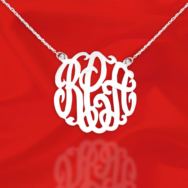 May include: A silver necklace with a large, round pendant featuring a monogram design. The pendant is made of a single piece of metal and has a delicate, intricate design.