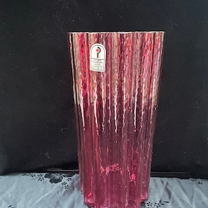 Puede incluir: Un jarrón de vidrio rosa alto con una superficie texturizada y ondulada. El jarrón tiene una etiqueta que dice "Pilgrim Cranberry Glass, Made in USA".