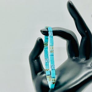 May include: A bracelet composed of small, square, turquoise-colored beads. The beads feature a mix of solid bright blue tones and marbled patterns with white and brown veining, strung together in a continuous, flexible circular band design.