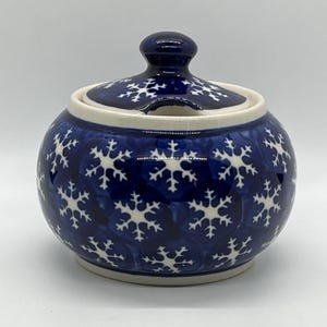 Op de afbeelding: Een ronde keramische suikerpot met deksel, voorzien van een diepblauwe achtergrond met een herhalend wit sneeuwvlokpatroon. De pot heeft een gladde, glanzende afwerking en een inkeping in de rand voor een lepel, met een bijpassend deksel.
