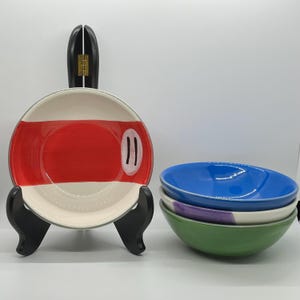 Könnte beinhalten: Ein Set aus vier Keramikschalen. Eine Schale steht auf einem schwarzen Ständer und zeigt ein cremefarbenes Inneres mit einem breiten horizontalen roten Streifen und zwei schwarzen vertikalen Linien. Die anderen drei Schalen sind blau, lila und grün.
