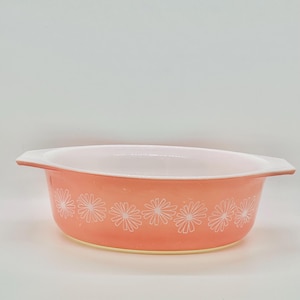 May include: A round, shallow casserole dish with two side handles. The exterior is a solid coral-orange color featuring a repeating pattern of white, thin-petaled daisy flowers encircling the base. The interior of the dish is smooth and white.