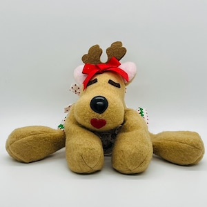 May include: A tan felt reindeer plush toy featuring brown antlers, a bright red bow, and pink inner ears. The reindeer has a black nose, a red heart-shaped mouth, and closed eyes. It wears a white apron with red polka dots and green trim.