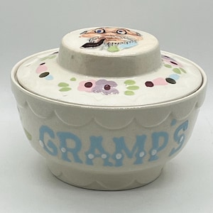 May include: A cream-colored ceramic bowl with a matching lid, featuring a scalloped edge design. The word GRAMP'S is painted in light blue on the front. The lid displays a hand-painted illustration of an older person with glasses and a pipe.