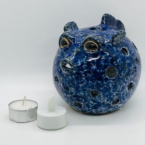 May include: A ceramic, blue pufferfish-shaped candle holder with black eye details and multiple circular openings. Two small, white candles sit beside the holder, one with a lit wick and the other with a battery-operated flame.