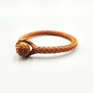 May include: A tan leather bracelet featuring a braided cord design and a spherical knot closure. The bracelet is constructed from a single continuous piece of woven leather that loops through the round knot to secure the circular shape.