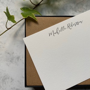 May include: A white stationery card with the name "Michelle Robertson" printed in black script. The card is partially inside a black box with a brown interior. Green ivy with white and yellow edges is in the background.