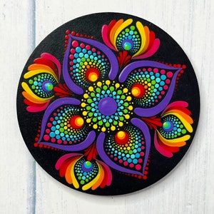 May include: A colourful mandala painted on a round wooden coaster. The mandala features a purple centre with concentric circles of green, yellow, blue, red, and orange dots. The mandala is surrounded by a black background.
