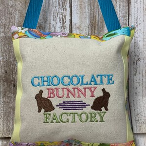 May include: A light yellow canvas pillow with a blue, pink, and green embroidered design that says "Chocolate Bunny Factory". The pillow has a blue ribbon attached to the top and a colorful patterned fabric border.