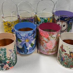 May include: A collection of decorative tin buckets with wire handles. Each bucket features a unique floral or nature-themed design, including butterflies, flowers, and an owl. The buckets have various rim colors, such as blue, pink, and purple.
