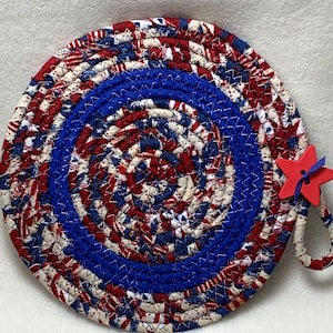 May include: A round, red, white, and blue fabric pot holder with a red star attached to the handle. The pot holder is made of braided fabric and has a patriotic theme.