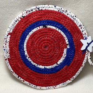 May include: Red, white, and blue circular woven trivet with a star-shaped tag attached. The trivet has a concentric circle design with white stars on a red background.