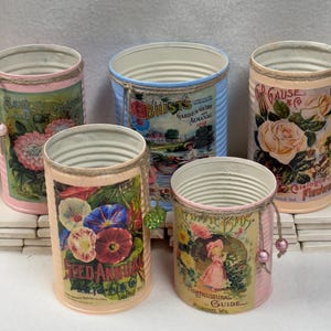 May include: Five repurposed tin cans, each adorned with vintage floral and botanical illustrations. The cans feature pink, blue, and peach backgrounds, with decorative twine and beaded accents. The cans are ideal for home decor or storage.