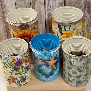 May include: Five decorative tin cans with various designs. One features a sunflower, another has a floral pattern with butterflies, and a third showcases a seashell theme. The remaining two cans have daisy and floral patterns. Each can is embellished with decorative trim.