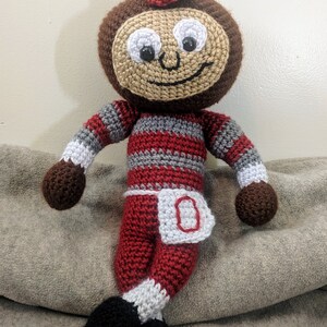 May include: A crocheted plush toy in the shape of a cartoon character wearing a red and white striped outfit with a white 'O' on the front. The toy has a brown face with black eyes and a smile.