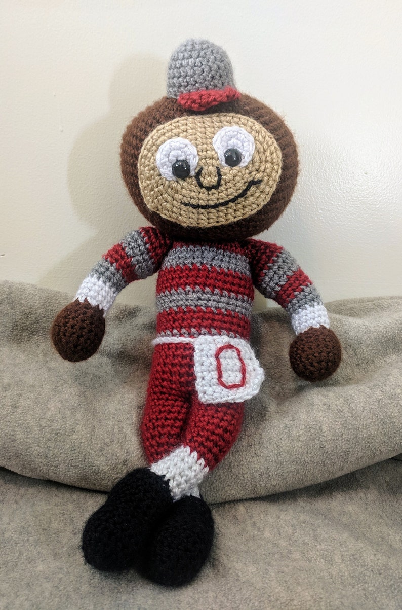 May include: A crocheted plush toy in the shape of a cartoon character wearing a red and white striped outfit with a white 'O' on the front. The toy has a brown face with black eyes and a smile.
