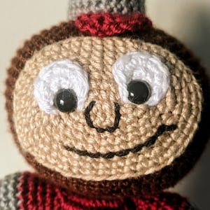 May include: A crocheted plush toy with a brown head, red and gray striped sweater, and a smiling face with black eyes and a white crocheted outline.