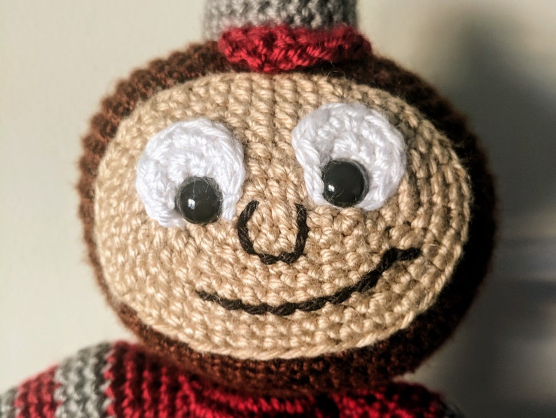 May include: A crocheted plush toy with a brown head, red and gray striped sweater, and a smiling face with black eyes and a white crocheted outline.