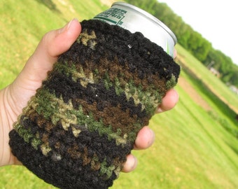 May include: A black, green, and brown crocheted can cozy with a camouflage pattern. The cozy is being held by a hand and is covering a can of soda.