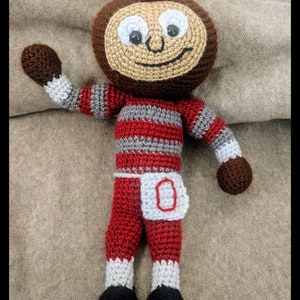 May include: A crocheted plush toy in the shape of a cartoon character wearing a red and gray striped shirt, red pants with a white 'O' on the front, and a gray hat. The toy is standing on a brown surface.