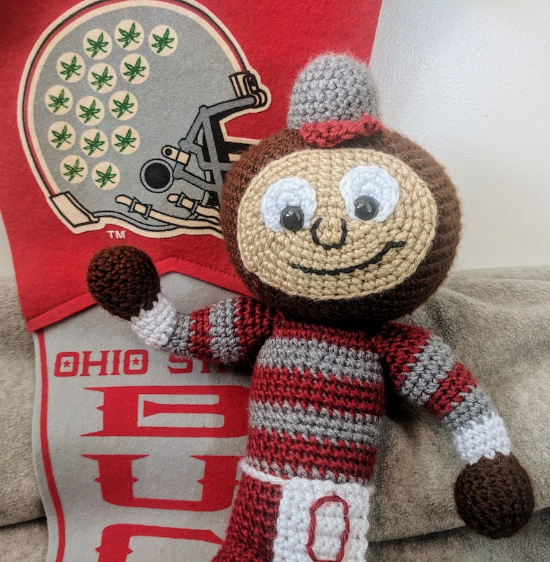 May include: A crocheted plush toy of Brutus Buckeye, the mascot of Ohio State University. The toy is wearing a red and gray striped outfit with a white 'O' on the front. The toy is holding its right arm up in the air.