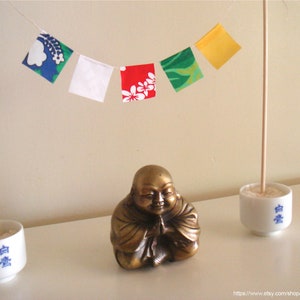 Pode incluir: Uma estátua dourada de Buda está posicionada sob uma série de bandeiras quadradas coloridas. Duas xícaras de cerâmica branca com caracteres japoneses azuis seguram palitos de madeira. As bandeiras apresentam padrões florais e abstratos.