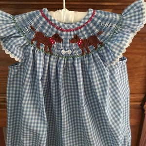 May include: A blue and white gingham baby dress with a smocked bodice featuring two brown dogs with red collars and a bone. The dress has short, ruffled sleeves.