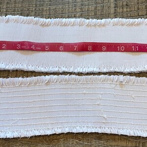 May include: Two white fabric strips with fringed edges are shown with a red measuring tape. The tape shows a length of approximately 14 inches. The top strip has a plain texture, while the bottom strip has horizontal lines.