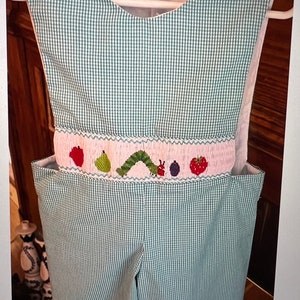 May include: Green and white gingham baby overalls with a pink embroidered band featuring a caterpillar, fruit, and berries.