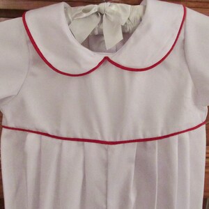 May include: A white baby onesie with red trim around the collar and the bottom. The onesie has a peter pan collar and short sleeves.