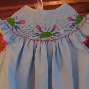 May include: Light blue baby dress with smocked bodice and crab embroidery. The dress features ruffled sleeves trimmed with pink pom-poms. The crabs are embroidered in green, pink, and blue. The dress is hanging on a white hanger.