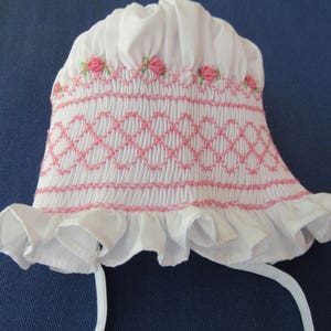 May include: White baby bonnet with pink embroidered details. The bonnet features a ruffled brim and a smocked design with pink diamond patterns. Small pink rose embroidery accents the top edge, and white ties are attached.