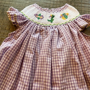 May include: A purple and white gingham dress with a smocked bodice and embroidered flowers. The dress has short, ruffled sleeves.