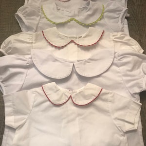 May include: Four white shirts with different colored trim on the collars. The collars are all Peter Pan style with scalloped edges. The trim colors are pink, green, red, and red.