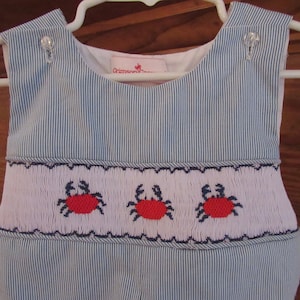 May include: A blue and white striped baby onesie with a white smocked panel featuring three red embroidered crabs. The onesie has a white collar and a label that reads "Crimson & Clover".