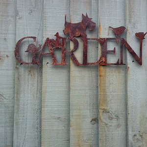 May include: Rusty metal sign with the word "GARDEN" in large letters. The sign is attached to a light gray wooden fence. The letters are decorated with small metal cutouts of animals and plants.