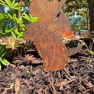 May include: A rusty metal angel with wings outstretched, standing in a bed of brown wood chips.