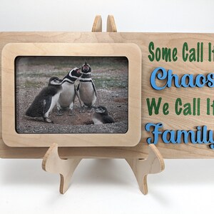 Puede incluir: Un marco de fotos de madera con un soporte, que presenta una foto de pingüinos. El marco tiene un recorte para la foto y el texto "Some Call It Chaos We Call It Family" en letras azules y verdes.