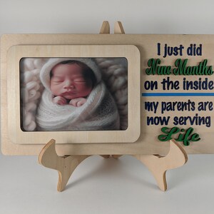 Puede incluir: Un marco de fotos de madera con una foto de un bebé recién nacido envuelto en una manta. El marco tiene el texto "I just did Nine Months on the inside, my parents are now serving Life."