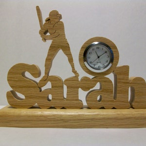 May include: Wooden clock with the name "Sarah" carved into it. A baseball player silhouette is standing on top of the name. The clock face is white with black numbers and hands.