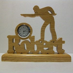 May include: Wooden clock with a silhouette of a person playing pool. The clock face is white with black numbers and hands. The name "Robert" is carved into the wood below the clock.
