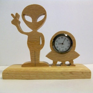 May include: Wooden clock with a carved alien figure and a flying saucer. The clock face is white with black numbers and hands.